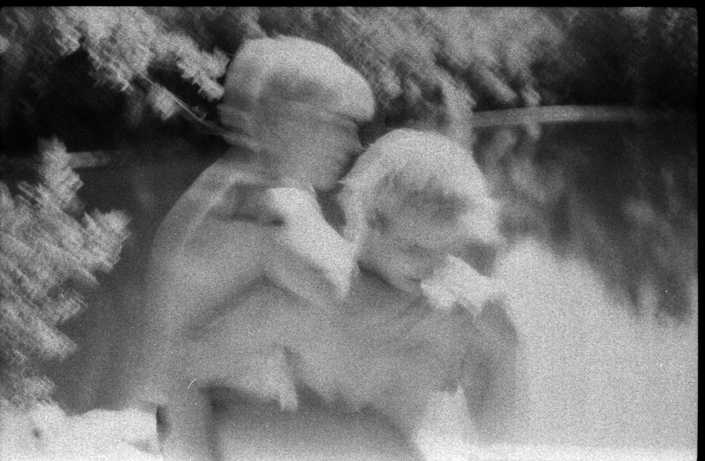 A blurry black and white image of two white people with blond hair. The person on the left has their arm around the other's neck. They are framed by foliage and in the background a body of water reflects the sky and trees.